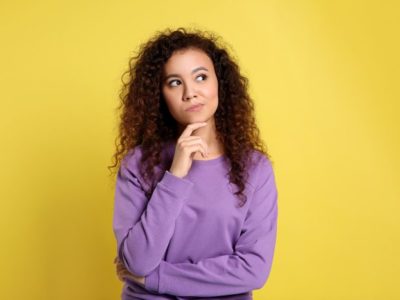 pretty young woman in a purple sweater standing against a yellow background making a questioning gesture with her hands and face - friendships