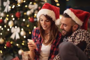 attractive young couple in Santa hats looking at a smart phone together in front of a lit Christmas tree - the holiday season