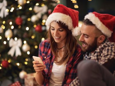 attractive young couple in Santa hats looking at a smart phone together in front of a lit Christmas tree - the holiday season