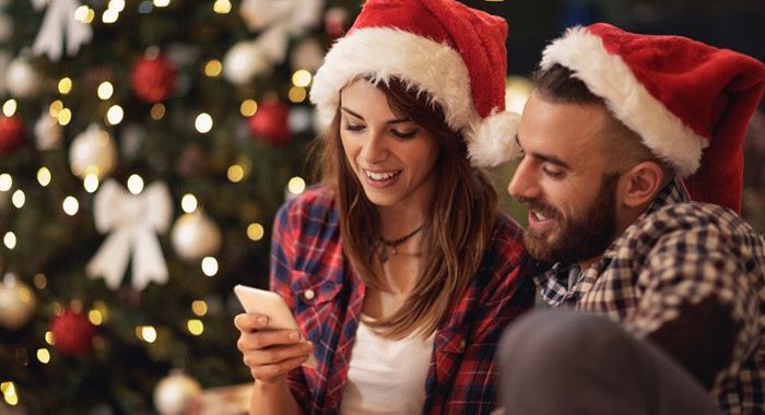 attractive young couple in Santa hats looking at a smart phone together in front of a lit Christmas tree - the holiday season