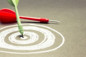 white hand drawn chalk target with real red and light green darts laying atop it - Goal-Setting Theory of Motivation