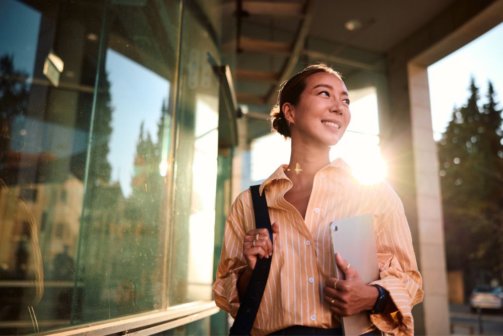 A woman in a striped shirt smiles while standing outside with a laptop and a bag, sunlight shining behind her and reflecting on a glass window.