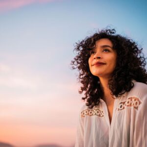 Woman with curly hair smiling peacefully while looking upward, standing outdoors at sunset under a pastel sky.