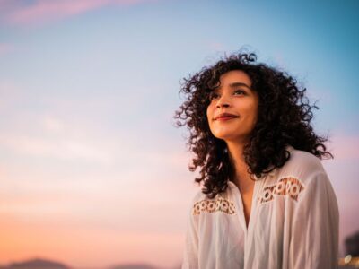 Woman with curly hair smiling peacefully while looking upward, standing outdoors at sunset under a pastel sky.