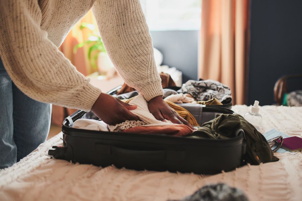 Person organizing clothes and personal items into an open suitcase on a bed in a softly lit room.