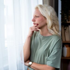 Woman standing by a window with a thoughtful expression, looking outside with her hand resting on her chin, appearing deep in thought or concern.