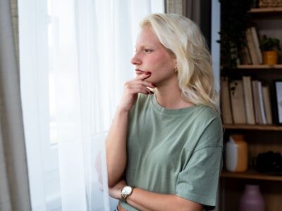 Woman standing by a window with a thoughtful expression, looking outside with her hand resting on her chin, appearing deep in thought or concern.
