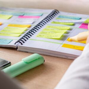 Close-up of a person highlighting tasks in a spiral planner filled with colorful sticky notes, with a phone and highlighter on the desk.