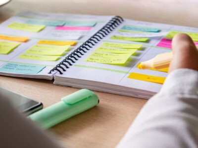 Close-up of a person highlighting tasks in a spiral planner filled with colorful sticky notes, with a phone and highlighter on the desk.