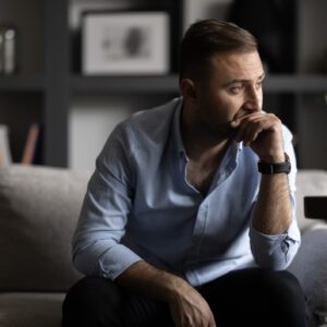 Man sitting on a couch, looking thoughtful and concerned, with his hand on his chin—suggesting inner struggle, reflection, or emotional distress.