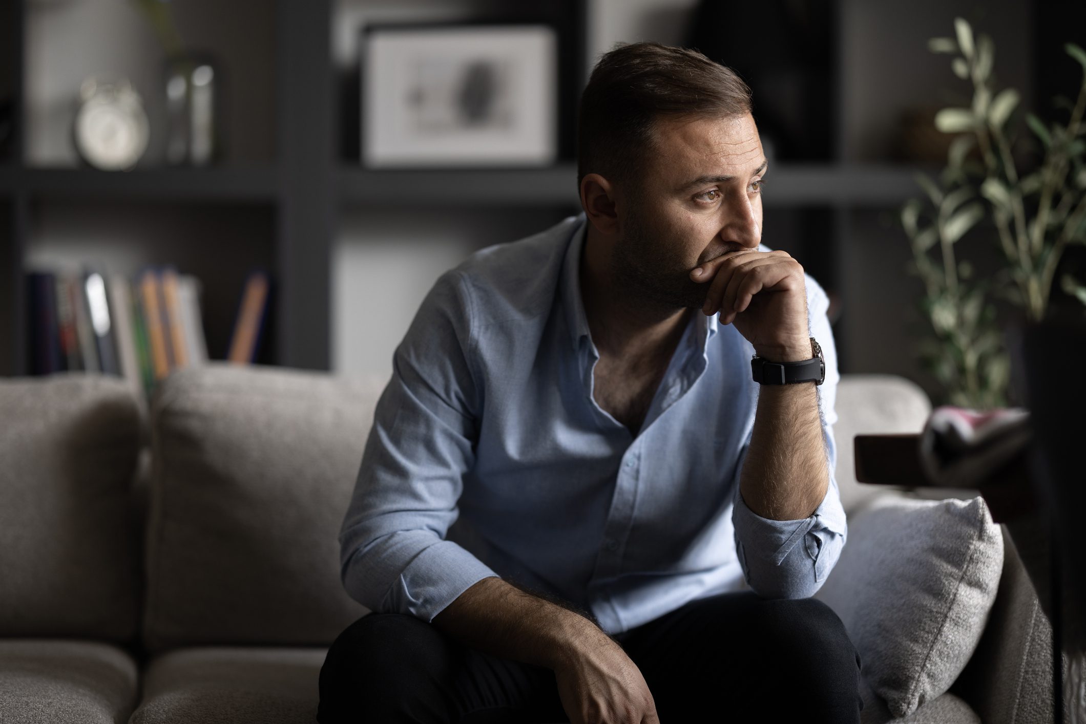 Man sitting on a couch, looking thoughtful and concerned, with his hand on his chin—suggesting inner struggle, reflection, or emotional distress.