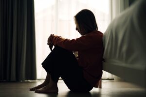 A woman sitting on the floor beside a bed, silhouetted against a bright window, with her head resting on her knees—suggesting sadness, reflection, or emotional pain.
