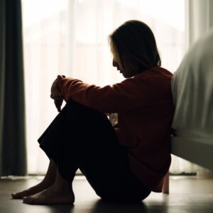A woman sitting on the floor beside a bed, silhouetted against a bright window, with her head resting on her knees—suggesting sadness, reflection, or emotional pain.