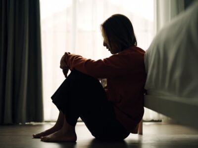A woman sitting on the floor beside a bed, silhouetted against a bright window, with her head resting on her knees—suggesting sadness, reflection, or emotional pain.