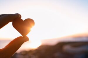 A hand holds up a small heart-shaped object against a bright sunset sky, with sunlight shining through. The scene feels peaceful and hopeful.