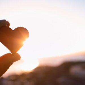 A hand holds up a small heart-shaped object against a bright sunset sky, with sunlight shining through. The scene feels peaceful and hopeful.