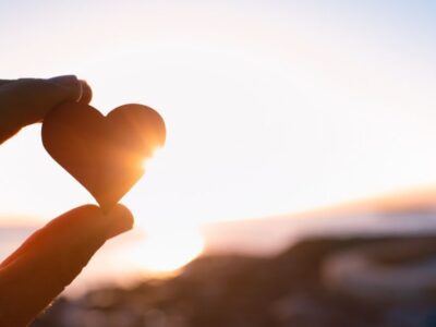 A hand holds up a small heart-shaped object against a bright sunset sky, with sunlight shining through. The scene feels peaceful and hopeful.
