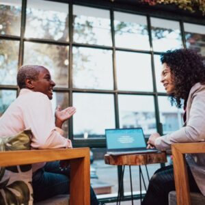 Two women sit across from each other in a warmly lit room, having a friendly conversation. One woman gestures while talking, and the other smiles as she listens. A laptop sits open on the table between them, suggesting a casual coaching or mentoring session.