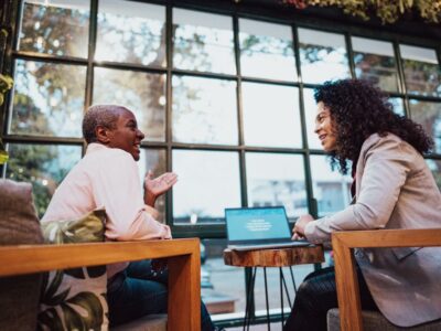 Two women sit across from each other in a warmly lit room, having a friendly conversation. One woman gestures while talking, and the other smiles as she listens. A laptop sits open on the table between them, suggesting a casual coaching or mentoring session.