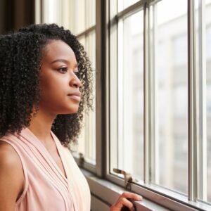 A thoughtful young woman stands by a window, gazing outside with a calm, reflective expression, symbolizing resilience and managing cravings in recovery.