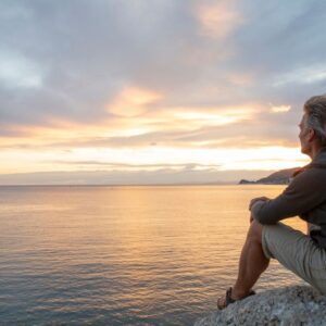 Man sitting on a rocky shore at sunset, gazing out over calm water with a thoughtful expression, suggesting reflection, peace, or solitude.