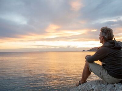 Man sitting on a rocky shore at sunset, gazing out over calm water with a thoughtful expression, suggesting reflection, peace, or solitude.