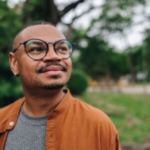 A man wearing glasses and an orange jacket stands outdoors, looking off into the distance with a thoughtful, content expression.