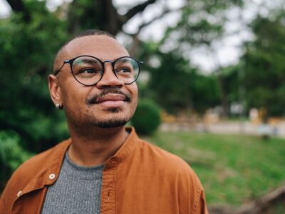 A man wearing glasses and an orange jacket stands outdoors, looking off into the distance with a thoughtful, content expression.