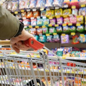 lose-up of a person pushing a shopping cart down a brightly lit grocery store aisle filled with colorful packaged sweets and snacks.