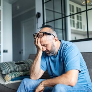 A middle-aged man sitting on a couch with his hand covering his face, looking stressed and exhausted, in a home setting.