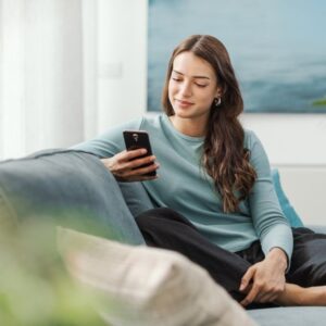 A woman sits comfortably on a couch, smiling as she looks at her smartphone.
