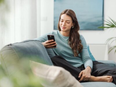A woman sits comfortably on a couch, smiling as she looks at her smartphone.