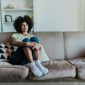 Woman sitting on a couch with arms wrapped around her knees, appearing deep in thought or emotionally reflective in a quiet room.