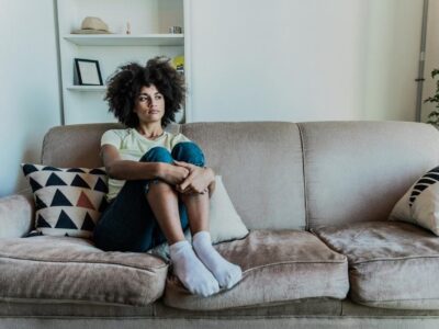 Woman sitting on a couch with arms wrapped around her knees, appearing deep in thought or emotionally reflective in a quiet room.