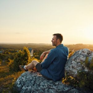 Man sitting on a rocky hillside at sunset, looking peaceful and reflective while enjoying a scenic view of nature.