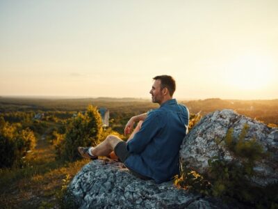Man sitting on a rocky hillside at sunset, looking peaceful and reflective while enjoying a scenic view of nature.