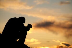 A person sitting outdoors at sunset with their head bowed and hands clasped, silhouetted against the sky, expressing stress or emotional struggle.