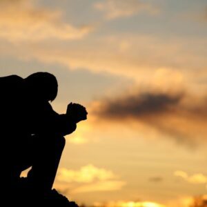 A person sitting outdoors at sunset with their head bowed and hands clasped, silhouetted against the sky, expressing stress or emotional struggle.