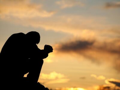 A person sitting outdoors at sunset with their head bowed and hands clasped, silhouetted against the sky, expressing stress or emotional struggle.