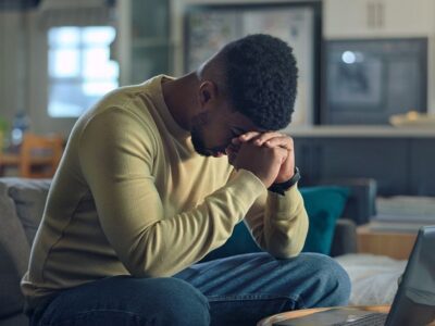 A man sits on a couch with his elbows on his knees and his hands clasped near his forehead, appearing stressed or deep in thought.