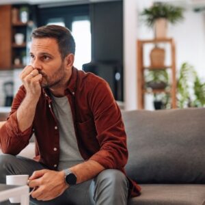 A man sitting on a couch holding a cup, looking thoughtful and anxious in a quiet living room.