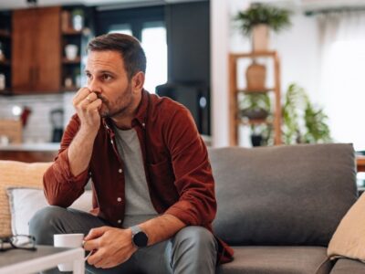 A man sitting on a couch holding a cup, looking thoughtful and anxious in a quiet living room.