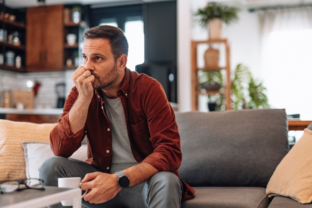 A man sitting on a couch holding a cup, looking thoughtful and anxious in a quiet living room.