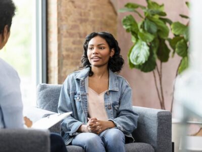 Waypoint - What’s Psilocybin-Assisted Psychotherapy For SUD. A young womans sits in a therapy session speaking with a therapist taking notes on a clipboard.