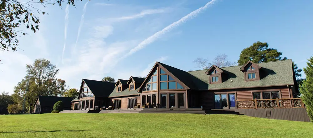 Large log cabin-style house with multiple peaked roofs, large windows, and green lawn in front, set against a clear blue sky with visible contrails and surrounding trees.