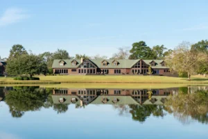 A large lodge-style building with green roofs is reflected in a calm pond, surrounded by trees and a manicured lawn under a clear blue sky.