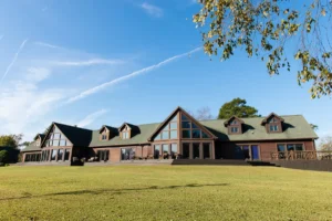 Large log cabin with many windows and dormers sits on a spacious, well-manicured lawn under a clear blue sky with some trees and a white contrail in the background.