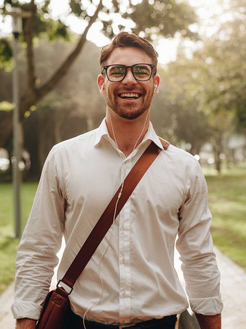 A smiling man with glasses, wearing a white shirt and carrying a shoulder bag, stands on a park path with trees and sunlight in the background.