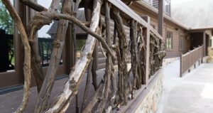 Close-up of a rustic porch railing made of twisted wooden branches attached to a brown log cabin, with stonework at the base and a walkway alongside the building.