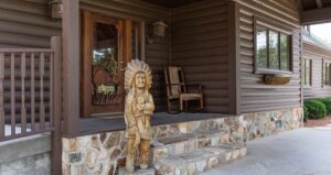 A wooden Native American statue stands by the steps outside a brown log cabin with a stone base, near a wooden rocking chair and a glass door with a decorative panel.